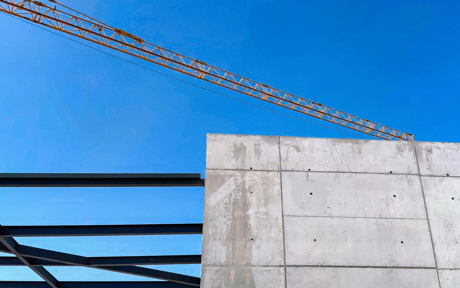 Industrial construction geometry showing raw concrete wall, steel framework, and crane against blue sky