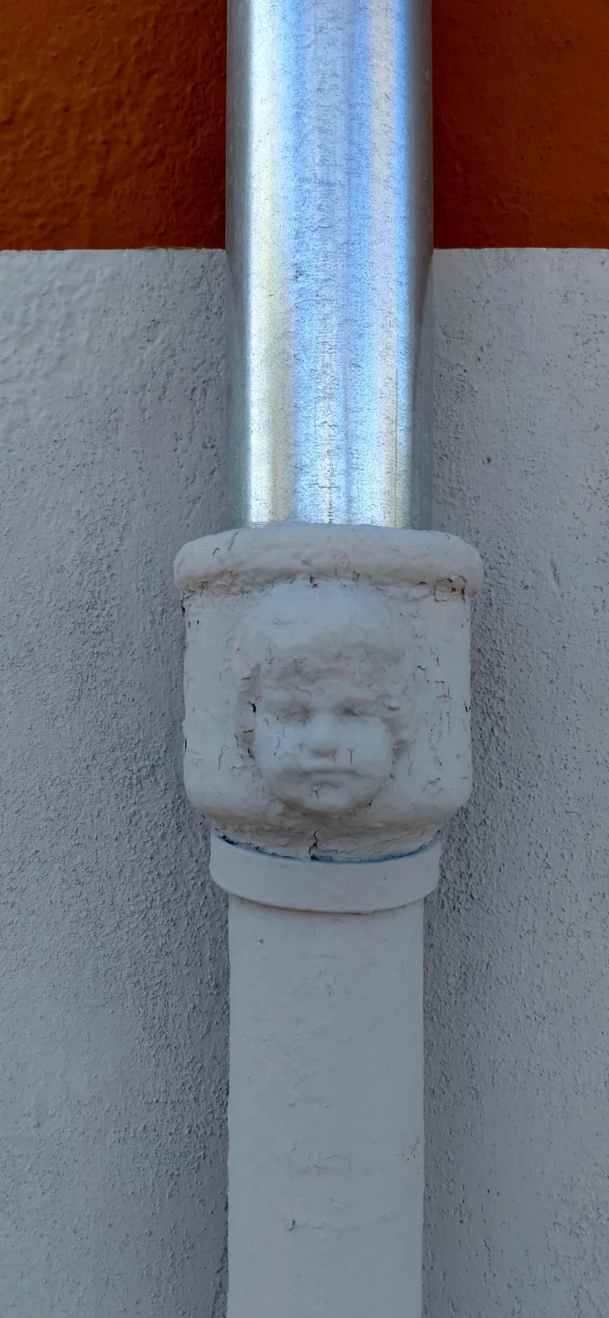 Architectural fixture detail of a carved face emblem on a white drainage pipe against a red wall