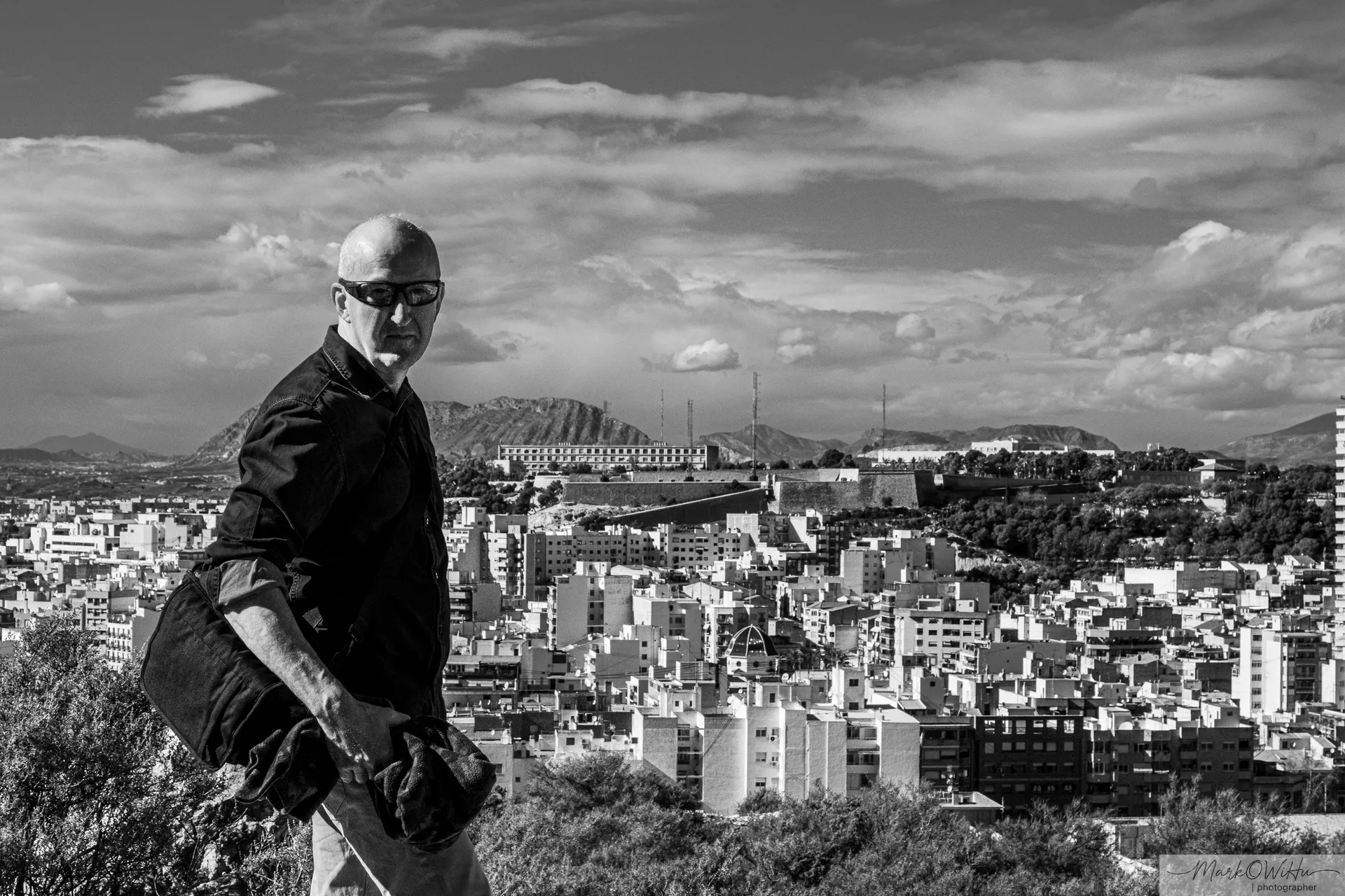 Black and white street portrait of a man with sunglasses and bag against sprawling city backdrop | Street & Humanity | Alicante province | MarkOWiHu Photographer Fotógrafo • markowihu.es