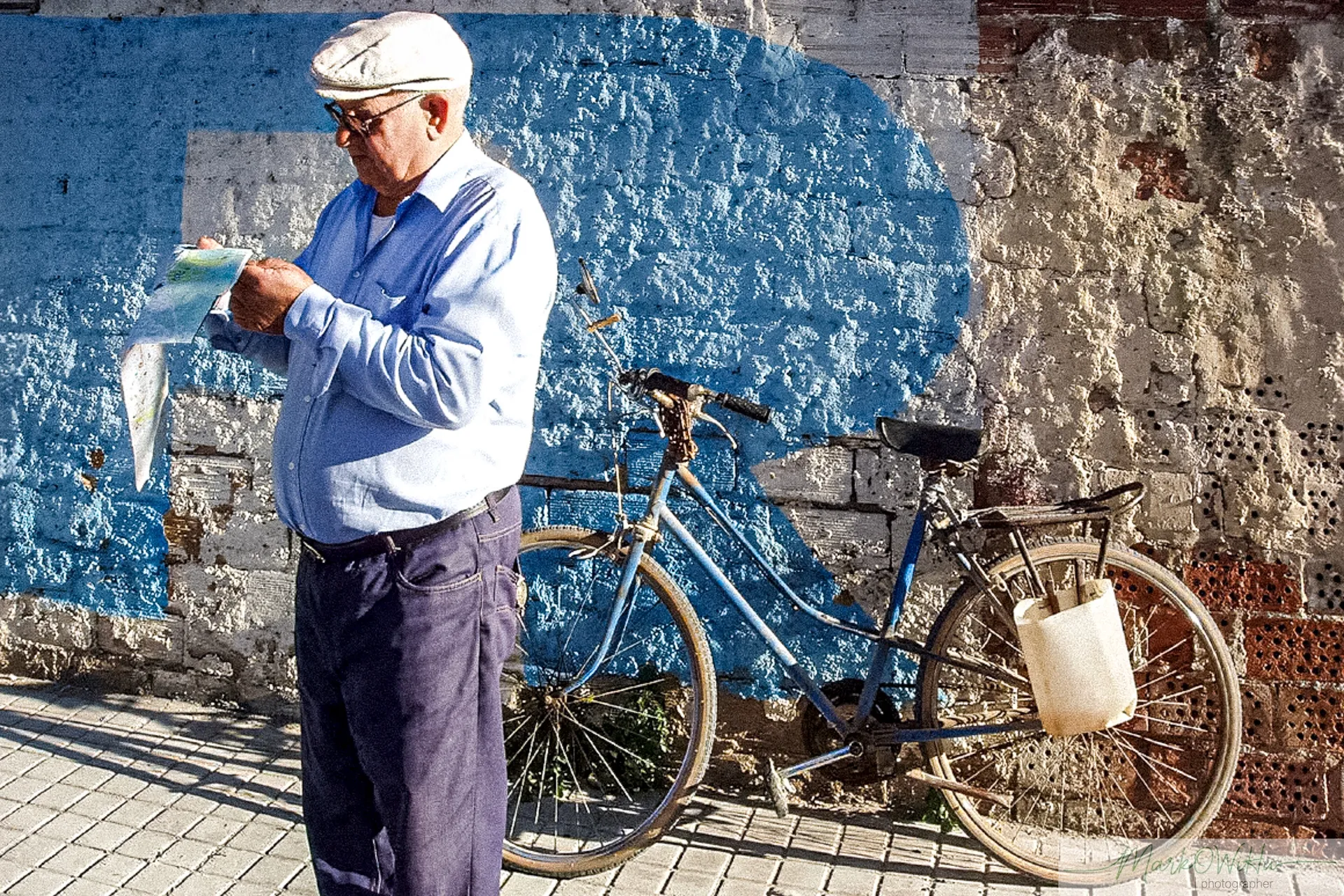 Candid street portrait of an elderly man reading beside his bicycle against a vibrant blue wall | Street & Humanity | Alicante province | MarkOWiHu Photographer Fotógrafo • markowihu.es