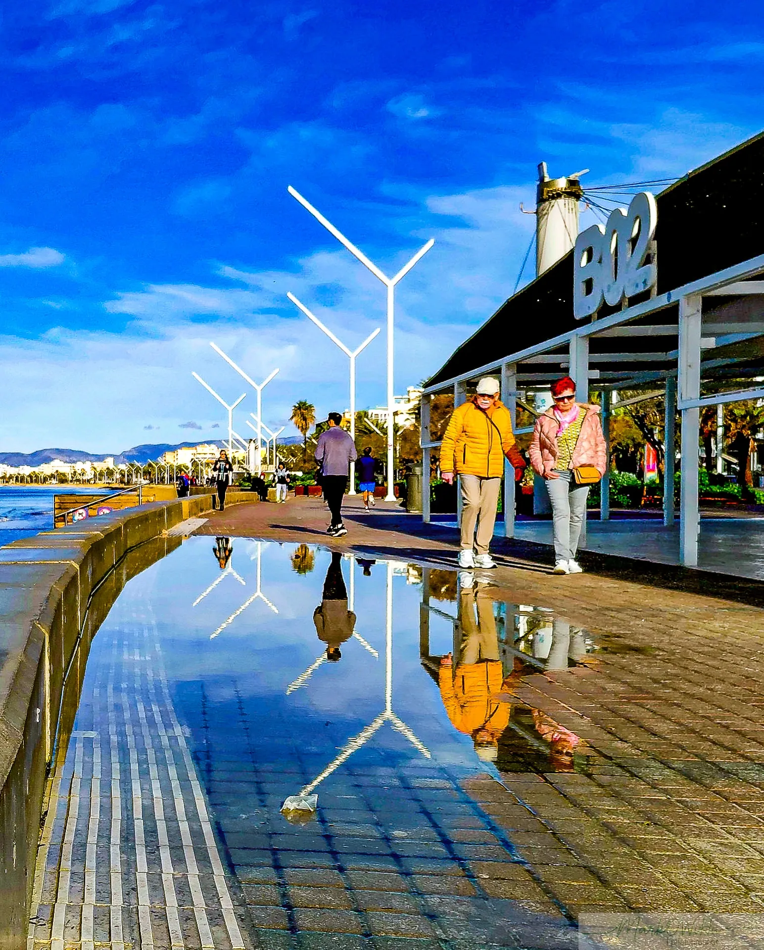 Saturated blue sky and puddle reflection of figures walking along modern seaside promenade | Street & Humanity | Alicante province | MarkOWiHu Photographer Fotógrafo • markowihu.es
