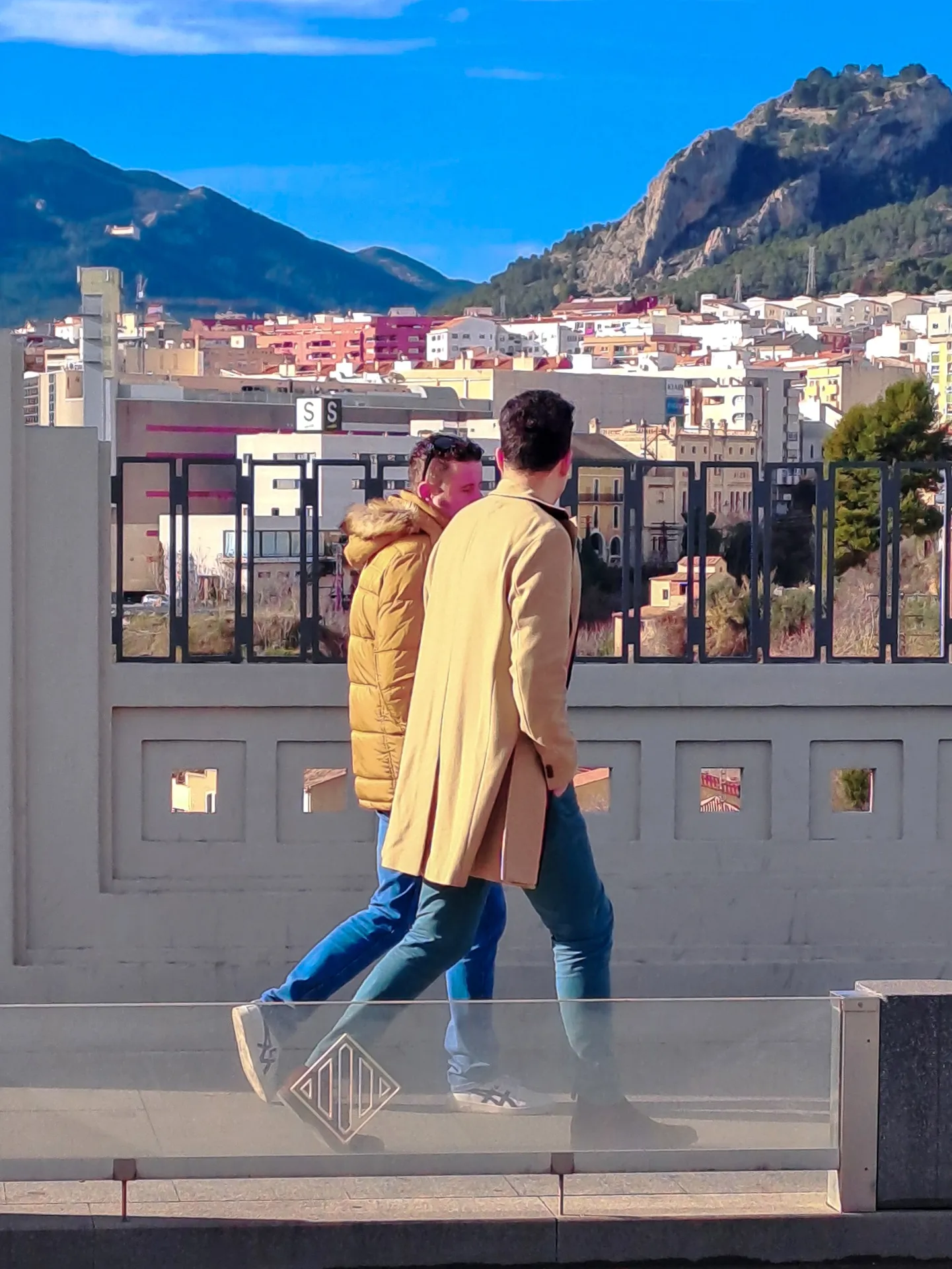 Urban commuter in bright yellow coat walking past glass railing with distant mountains | Street & Humanity | Alicante province | MarkOWiHu Photographer Fotógrafo • markowihu.es