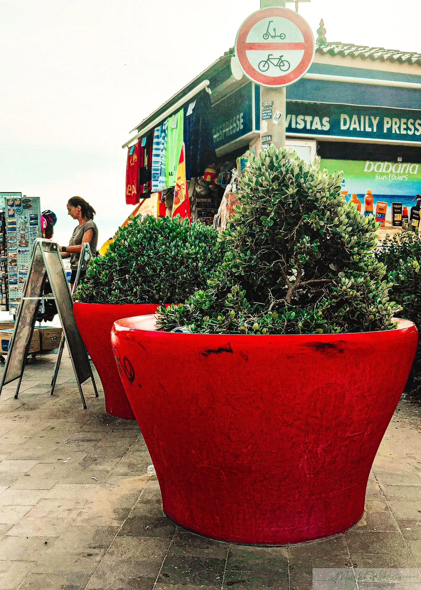 Vivid red planter pots in foreground leading to resort souvenir press kiosk and street scene | Street & Humanity | Alicante province | MarkOWiHu Photographer Fotógrafo • markowihu.es
